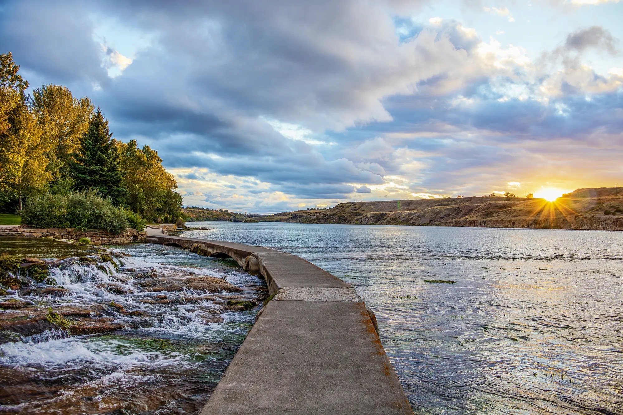 A scenic view of a Montana river at sunset with hills and trees in the background, promoting peaceful living in Cascade Communities.