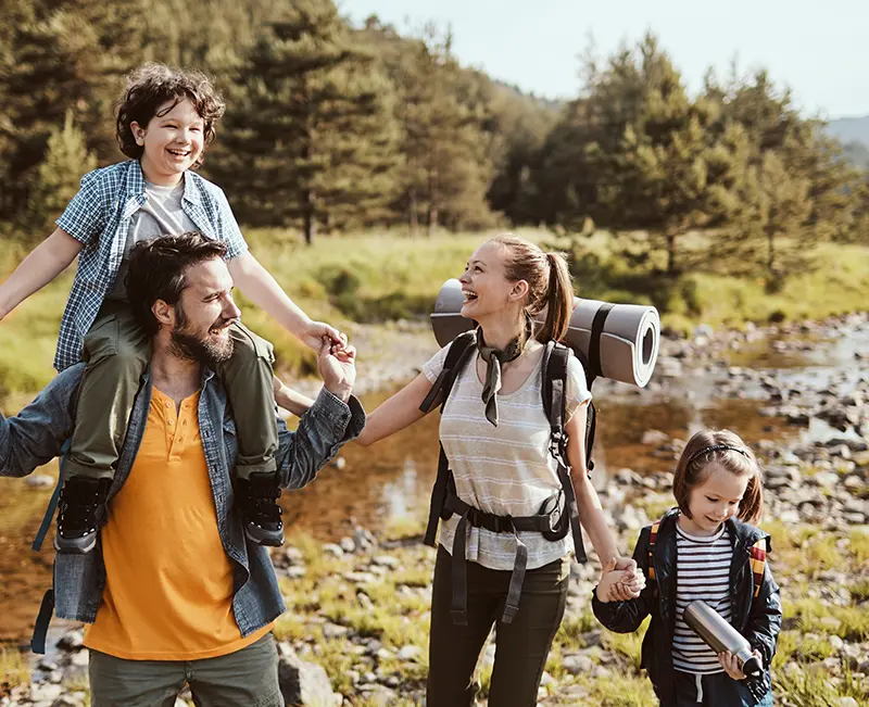 A smiling family with children and backpacks hiking outdoors, representing the active lifestyle offered at Cascade Communities in Great Falls, Montana.